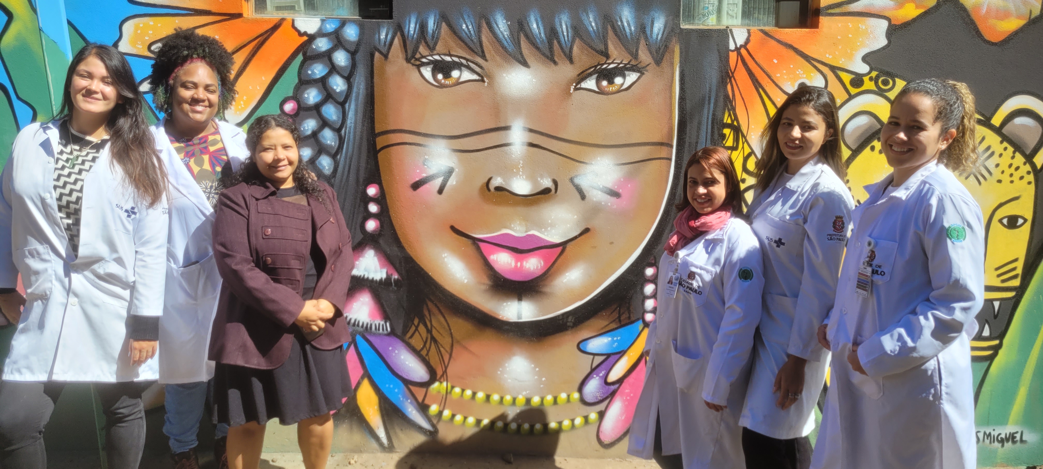 Na imagem, aparecem cinco mulheres com jaleco branco que integram a equipe de profissionais da UBSI do Jaraguá. Junto a elas está Poty Poran Turiba Carlos, gestora da UBSI. Ao fundo há um painel pintado com figuras coloridas de flores, uma onça e uma jovem indígena ao centro